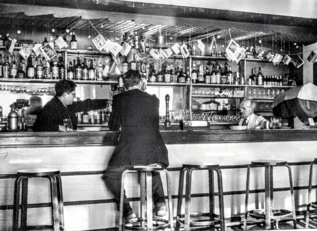 A man in a suit sits at the bar while Frank directs his assistant.
