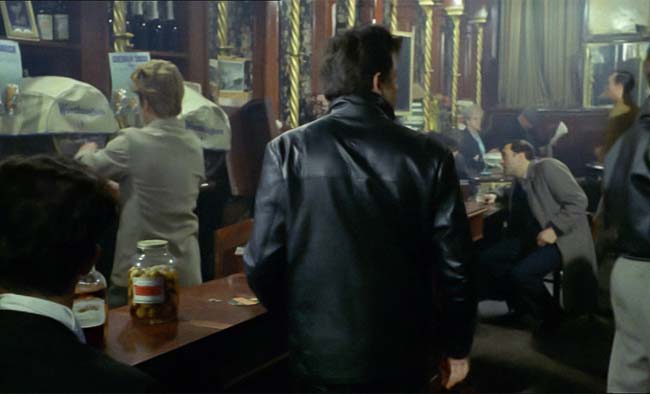 A man in a leather jacket stands next to a jar of pickled eggs in a crowded pub.