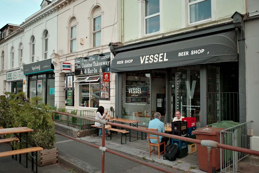 The exterior of Vessel with a dark grey awning and tables outside.