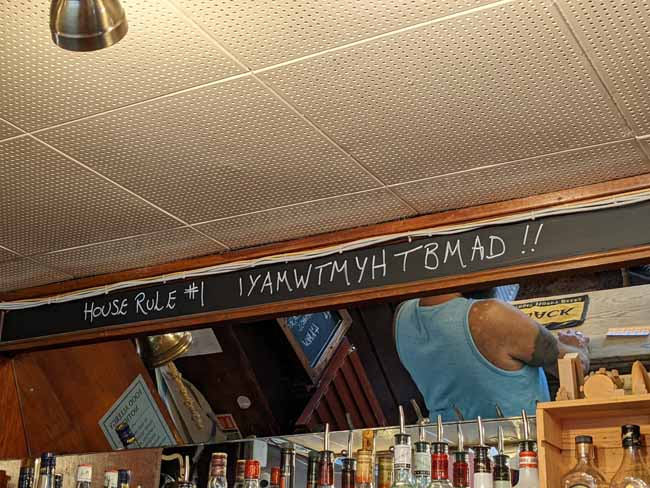 A view of the mirror behind a bar with the barman's shoulders reflected in it. In chalk above is written the cryptic message "Rule number 1: IYAMWTMYHTBMAD"