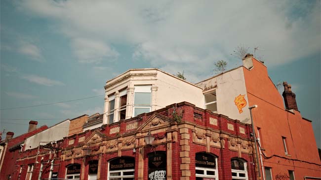 A decaying Victorian pub building with weeds growing from its roof and graffiti on its wall.
